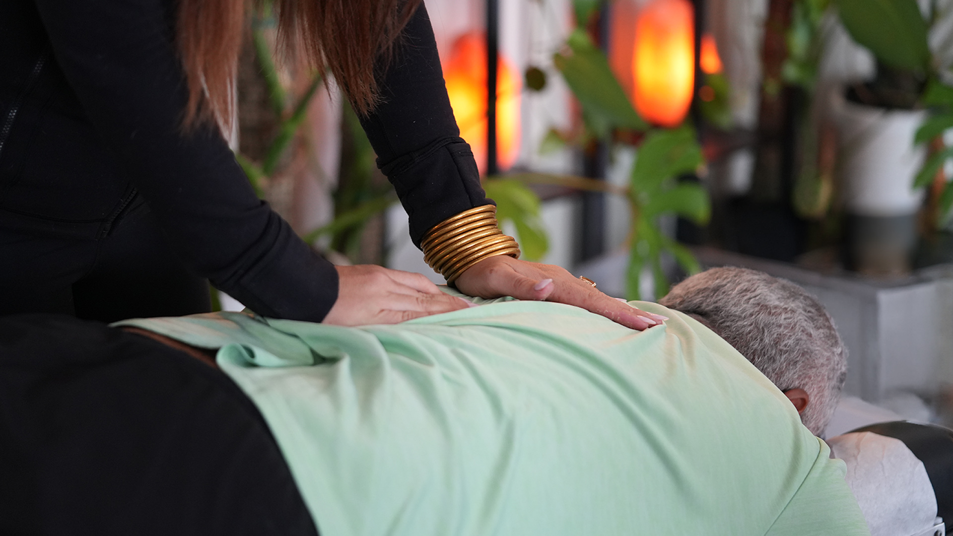 Therapist pressing on a person’s back during a back massage; the therapist wears multiple gold bangles on one wrist, with a green sheet and plant-filled background.