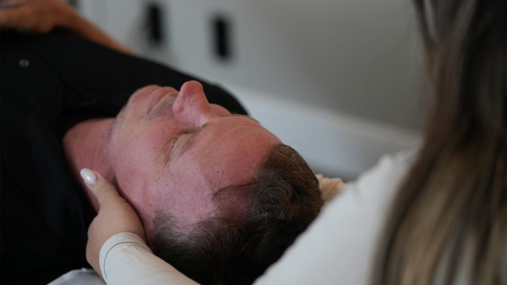 Caring chiropractor services - Express Life Chiropractic | Chiropractor Fort Lauderdale. Older man lying on his back with a flushed face, supported by a caregiver holding his head near the pillow(s). Best guess of medical setting or exam.