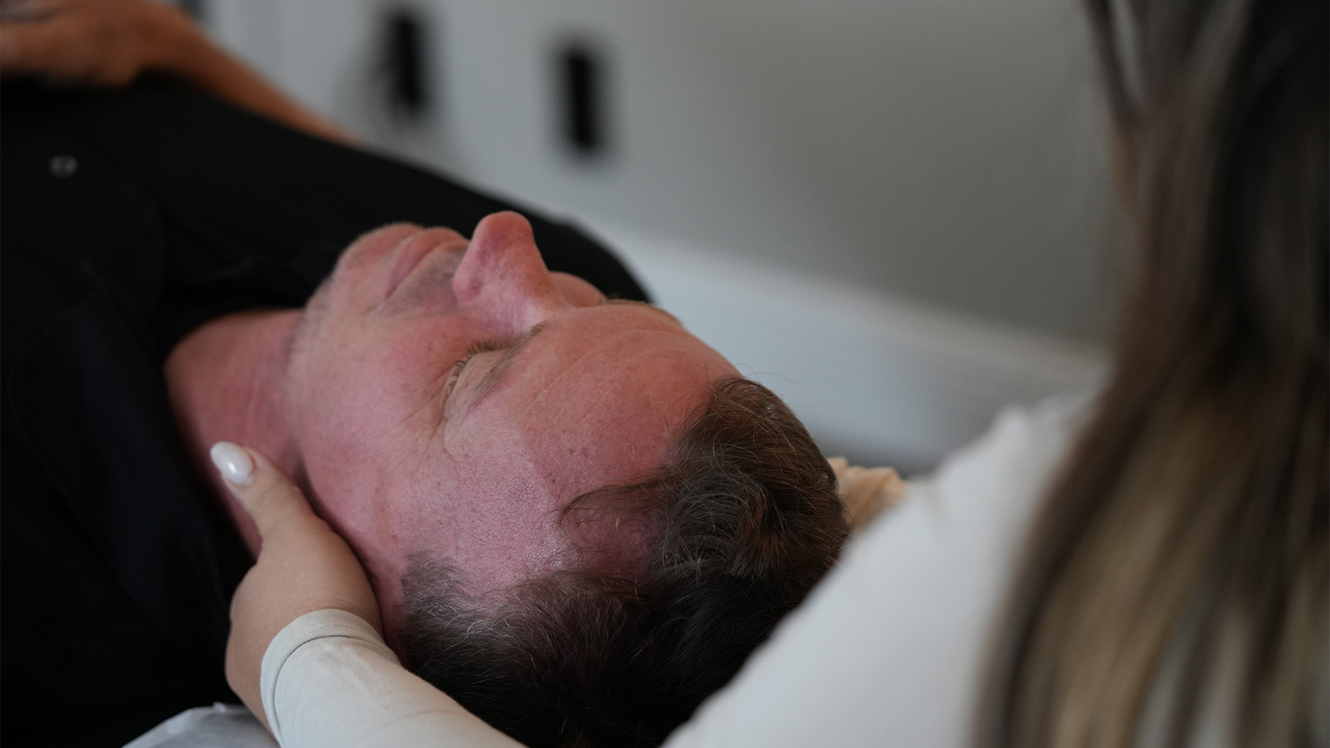 Older man lying on his back with a flushed face, supported by a caregiver holding his head near the pillow(s). Best guess of medical setting or exam.