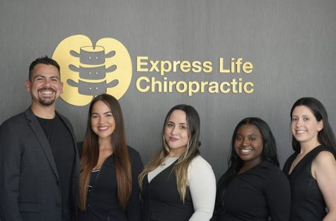 Five smiling staff members posing in front of a gray wall with the Express Life Chiropractic gold logo and sign.