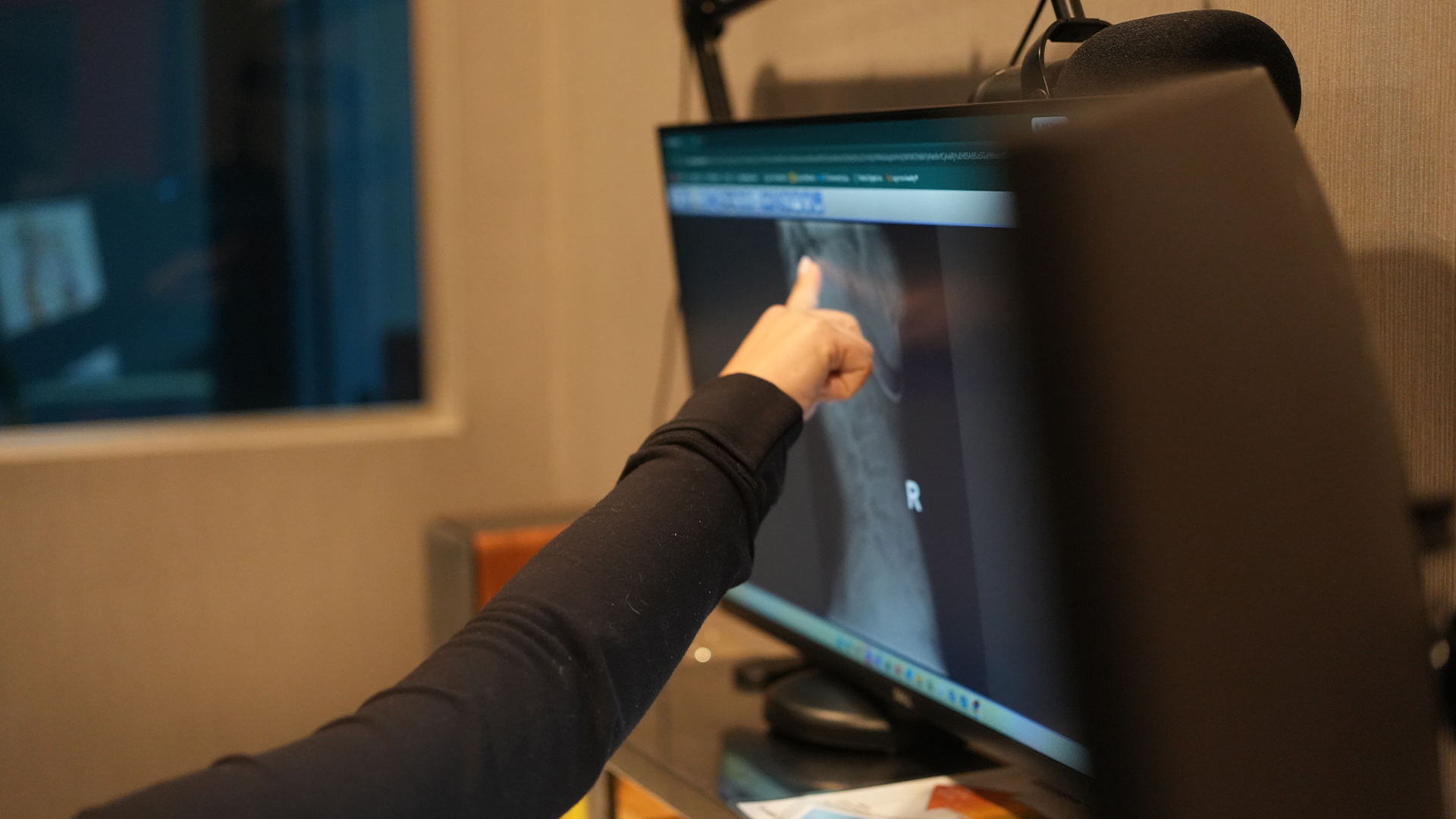 Arm in a black sleeve points at a computer monitor in a dim office or studio, mic arm visible nearby.