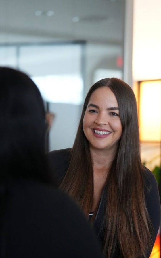 Smiling woman with long brown hair talking to someone in a bright office.