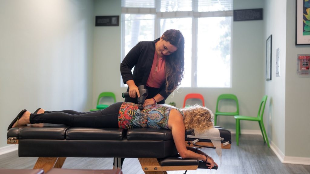 Therapist uses a handheld treatment wand on a patient lying face down on a massage/table in a bright clinic, with colorful chairs in the background.