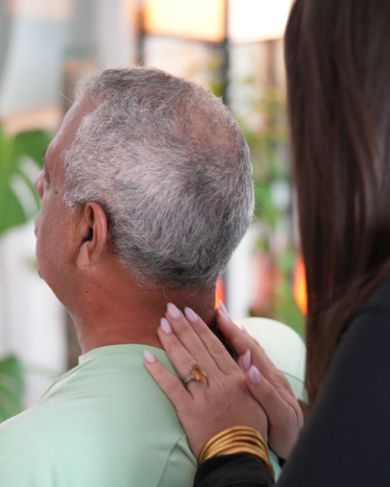Older man with gray hair wearing a mint shirt, as a woman's hand rests on his neck in a comforting gesture