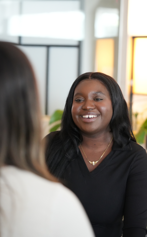 Smiling Black woman in a black top with a gold necklace, talking to a coworker in a bright office.