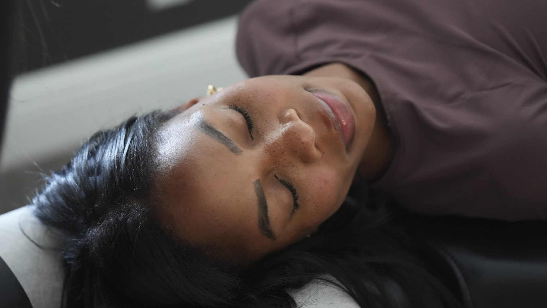 Woman with dark hair lying down on her back with eyes closed, wearing a maroon top.